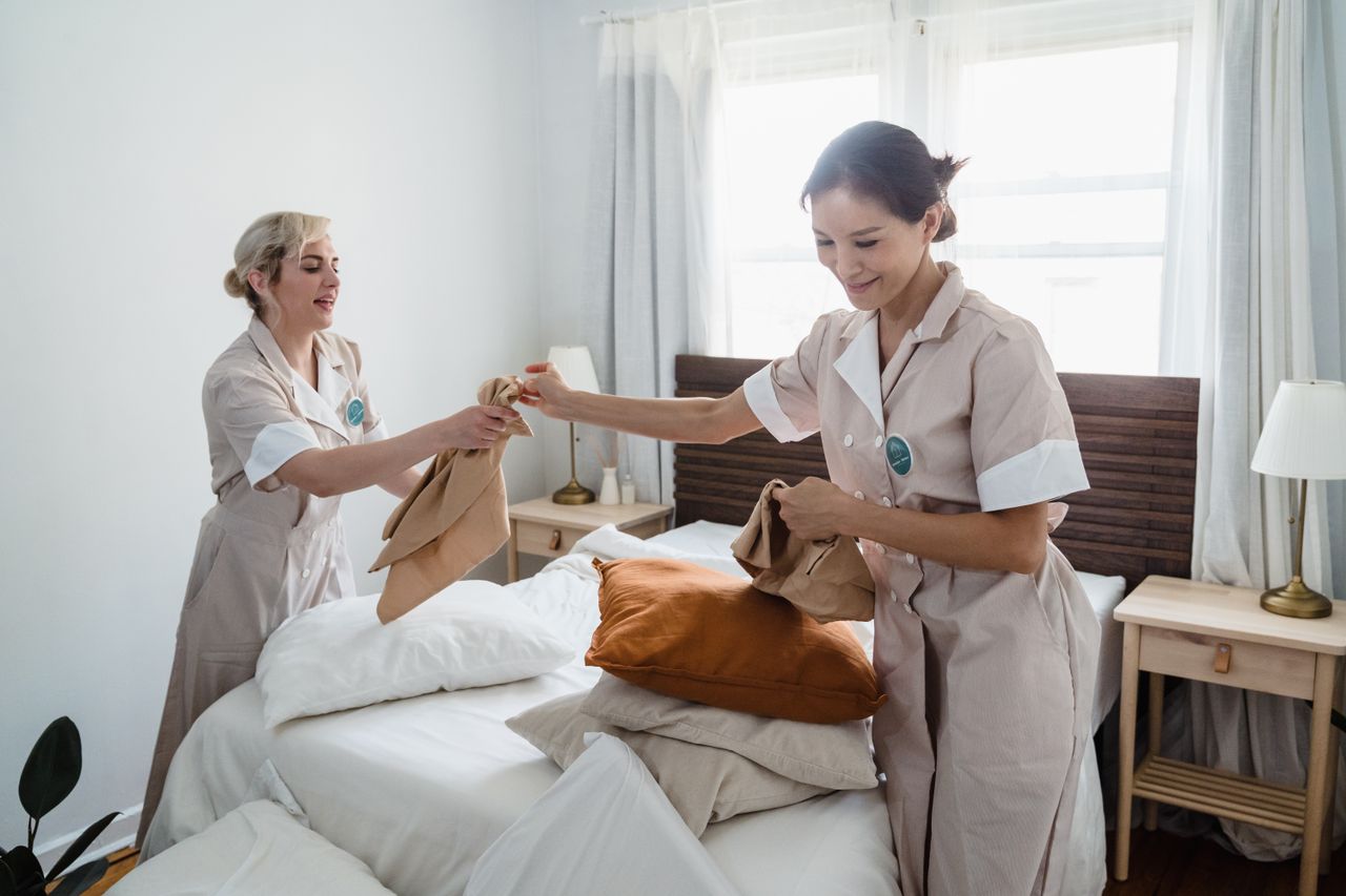 Two female housekeepers in uniform efficiently prepare a bed in a bright, well-lit room.